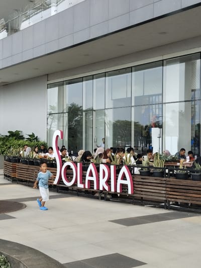 A young boy in a blue shirt and white shorts is running toward a large red sign that reads "SOLARIA" in front of a restaurant in Telukjambe, Indonesia. The restaurant is located at coordinates -6.301 latitude and 107.279 longitude. There are numerous people seated at tables inside the restaurant, some are interacting with each other while others are looking at the sign. The boy is alone and appears to be excited. There are numerous potted plants lined up along the front of the restaurant, and the floor is made of grey tiles. The restaurant is located inside of a modern building with a grey exterior. The windows are large and the door is glass. There is a small amount of greenery in front of the building.