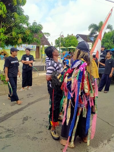 The image depicts a scene on a paved street in front of a building in Kecamatan Kedawung, Indonesia, located at latitude -7.52 and longitude 111.029. There are four men and one woman in the scene. Three men are wearing black trousers, two are wearing black shirts with one of them having a black and white striped shirt layered on top. The fourth man is wearing a black shirt covered in colorful ribbons and has a black bandana on his head. The woman is wearing a black t-shirt and black trousers, and is holding a hoop in her right hand. Two men in black shirts with black trousers stand to the left, one of them is looking at a man to the right, and the other is looking directly at the camera. The man in the striped shirt is facing the man in the black bandana and looking at him while gesturing towards the man in the black bandana. The man in the black bandana is looking off to the side, his head tilted up slightly. The woman in the scene is walking towards the right, her feet slightly off the ground. The man in the black bandana and the man with the black and white striped shirt are standing in the foreground facing each other. Two men are standing behind them looking towards them. The man in the black bandana has a large bundle of colorful ribbons tied around his waist and hanging to the ground. There is a white and red flag on a pole behind the men, and a building to the right of the men. The building appears to be a school, with a banner on the front. There is a tree to the left of the men and a building in the background to the right of the flag. The environment is a sunny, open-air setting with a paved road.