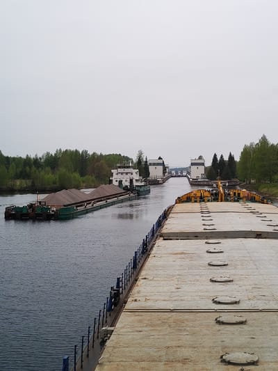 The image depicts a waterway, possibly a canal or wide river, near Alekseevskoe, Russia. In the foreground is a large, flat-decked barge, its surface exhibiting a repetitive pattern of circular access covers. A blue railing runs along the port side of the barge. In the water to the left, a smaller, loaded barge being towed by a tugboat is heading away from the viewer towards a double-chambered canal lock system in the distance. The lock chambers are marked with the number "5" on their exteriors. The surrounding landscape consists of green trees lining the banks of the waterway. The sky is overcast, creating a diffused light.