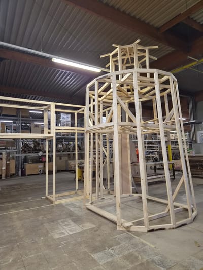 The image shows a large, unfinished wooden frame structure, likely a set piece or a large-scale art installation, under construction in what appears to be a workshop or warehouse. The structure is made of light-colored lumber, forming a complex, multi-layered framework with geometric shapes and open spaces. It has a somewhat organic or stylized form, with a rounded section at the top and a more rectangular base.
The setting is industrial, with a concrete floor marked with lines and some scuff marks. The ceiling is high, with visible beams and corrugated metal roofing. Fluorescent lighting fixtures are present, suggesting an indoor environment. In the background, there are shelves stocked with various objects, including boxes, equipment, and what looks like industrial machinery. A yellow object, possibly a piece of equipment, is visible further back.
The image is oriented vertically, with the structure filling most of the frame. The lighting suggests daytime, but the specific time of day is not discernible. There are no people visible in the image, indicating that the work might be paused or the construction is in a phase where human presence isn't captured. The overall impression is one of a large project in progress within a functional, working environment. There is a box with the number "4" visible in the background.