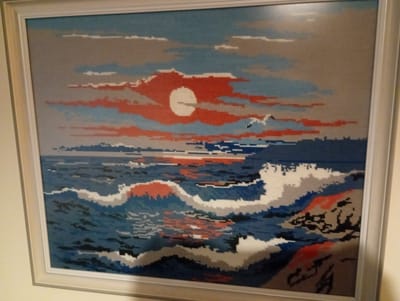 This is a framed cross-stitch artwork depicting a seascape at sunset. The sky is filled with wispy clouds in shades of gray, blue, and vibrant red, with a bright orange sun partially obscured by the clouds. A white bird is visible in the sky to the right of the sun. The sea below is rendered in shades of deep blue and white, with large waves crashing, indicated by white foam. Rocky landmasses, colored in gray, black, and red, are visible on the right side of the image. The artwork is framed in a light-colored frame. The scene is a representation of nature and does not contain any recognizable landmarks or elements that would specifically place it within Kajaani, Finland.