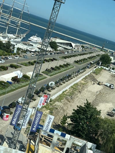 The image shows a waterfront area in Gdynia, Poland, with a large sailing ship docked in the harbor. A modern white ferry is also visible. In the foreground, a construction crane is positioned above a building under construction. Several flags, including "BAUHAUS" and "ATLAS WARD," are flying near the construction site.

The background features a promenade with trees and parked cars, leading to the sea. The sky is clear and blue. The scene appears to be a sunny day. The construction site itself shows excavated earth and some construction equipment, including a mini excavator and a white van. There are also tents set up near the promenade. The overall impression is a blend of maritime activity, ongoing development, and public space.