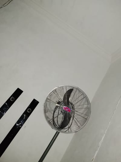 The image shows a floor fan in a room. The fan is silver and has a red and blue logo on it. There are two black, reflective bars in the foreground, angled diagonally from the bottom left. The walls and ceiling are a light color and appear to be textured. There is a slight shadow on the ceiling. The scene is indoors. The location is Ozuri, Nigeria.