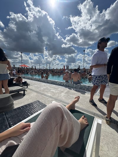 This image depicts a crowded outdoor rooftop pool facility in Gainesville, Florida, USA, under a bright, sunny sky with numerous scattered cumulus clouds.

In the immediate foreground, from a first-person perspective, the lower torso, legs, and bare feet of an individual are visible, resting on a dark-colored lounge chair. This individual wears light-colored, vertically striped wide-leg pants. Their left hand is visible, holding a dark smartphone. Their toenails are painted dark.

In the mid-ground, to the right, a male figure stands facing right, wearing a white short-sleeved t-shirt, dark baseball cap, sunglasses, and blue and white pineapple-patterned swim shorts. He wears dark athletic shoes with white soles and a bracelet on his left wrist. To his right, partially obscured, is another male figure wearing a black t-shirt, light shorts, and dark shoes. To the left mid-ground, a female figure stands facing away from the camera, wearing a white off-the-shoulder top and light blue denim shorts. Her dark hair falls down her back. Behind her, a partially visible person reclines on a dark lounge chair.

The background is dominated by a rectangular swimming pool filled with a large gathering of people, estimated to be over fifty individuals, primarily young adults. Most are partially submerged in the water, socializing. The pool edge features light-colored tiling. Around the pool perimeter, several tall, dark light poles and striped umbrellas (red and black-and-white) are visible. The bright sun is positioned high in the sky, causing a strong glare in the upper center of the image. The overall environment suggests a lively daytime social event.