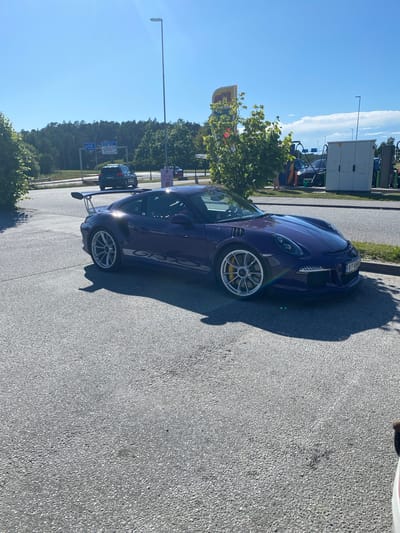 This media file depicts a dark purple Porsche 911 GT3 RS parked on an asphalt surface, likely at a rest stop or service area, given the presence of a sign with a Shell logo and a building that appears to be part of a facility with doors. The car is positioned at an angle, with its front left wheel visible and a large spoiler extending from the rear. The license plate reads "BT 494".

In the background, there is a tree with green foliage, other vehicles, and signage indicating directions to "E18 300m" and "Oslo 24km." The sky is clear and bright blue, suggesting a sunny day. The shadow of the car and possibly a person are cast on the ground, indicating it is daytime. The overall scene conveys a sense of a brief stop during a journey, likely in a scenic area given the trees and open sky. The car's sporty design and the presence of a highway sign suggest it is being driven on a road trip or used for recreational driving. The image does not capture any people, but the presence of a car and a road implies human activity. The weather appears to be pleasant, with no signs of rain or clouds.