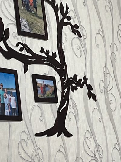 The image shows a dark brown tree-shaped wall decal with multiple picture frames attached to its branches. Three framed photographs are visible. The top frame contains a photograph of people standing on grass with a rocky area in the background. The frame on the left side contains a photograph of several people standing outdoors, with a car and a blue sky visible. The frame on the right side shows a photograph of two people standing by a body of water. The decal is mounted on a wall with a textured wallpaper featuring a pattern of light gray swirls. The scene is indoors, likely a home. There is no information in the image to suggest it is located in the city of Serov, Russia.