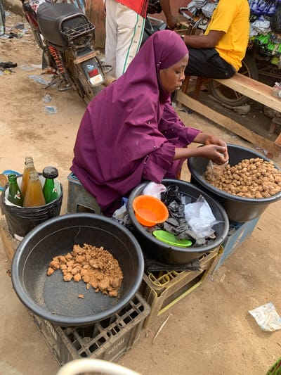 The image depicts a street scene in what appears to be a marketplace in Kafanchan B, Nigeria. A woman wearing a purple hijab is seated beside three large black plastic tubs, filled with what appear to be edible goods. One tub contains tan, roughly spherical objects, possibly fried dough or nuts. A second tub holds a mix of items, including orange and green plastic containers, along with clear and black plastic bags. The third tub contains a brown, lumpy substance, possibly a type of bean or seed. The woman appears to be handling the food, possibly portioning it for sale. Beside the tubs is a stack of empty plastic crates and a black plastic bag holding several glass bottles, possibly soft drinks or other beverages. A man in a yellow shirt is partially visible in the upper left, sitting on a low wooden structure. In the lower left, part of a motorbike is visible, suggesting transport to and from the market. The ground is dirt and scattered with debris.