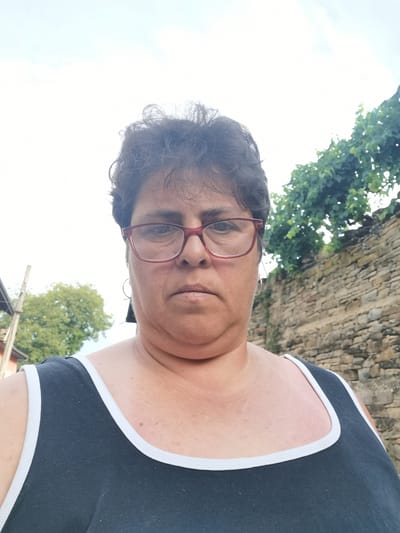 The image is a selfie taken outdoors. A middle-aged woman with dark, curly hair and glasses is the central subject. She is wearing a dark tank top with white trim. Behind her, a stone wall and lush green foliage are visible, suggesting an outdoor environment, possibly in a rural or semi-rural setting. To the left, a wooden pole and some structures indicate a residential area. The background is bright and overcast, suggesting daytime. The woman's expression is neutral, and she is looking directly at the camera. The photo is taken from the chest up. Lomets is a village in Northern Bulgaria, known for its natural beauty and traditional architecture. This setting aligns with the visual cues in the image.