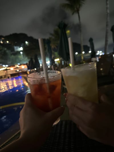 The image, rotated 90 degrees counter-clockwise, captures a nighttime scene of two individuals holding drinks in an outdoor setting in Rio de Janeiro, Brazil. Two hands are prominent in the foreground, each grasping a glass. The left hand holds a tall glass containing a light yellow, possibly icy or blended beverage with a white straw. The right hand holds a glass with a vibrant red or orange drink, also featuring a white straw and condensation on the glass, suggesting it is cold.
In the blurred background, the dark night sky is dotted with numerous soft, glowing artificial lights, creating a warm and inviting atmosphere. Silhouetted palm trees are visible, indicating a tropical location. Below the drinks, reflections of lights shimmer on a body of water, which appears to be a swimming pool, adding to the resort-like ambiance. The scene conveys a sense of leisure and enjoyment during an evening out. No text is visible in the image.