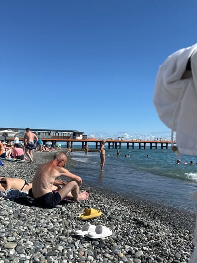 The image depicts a pebble beach on a sunny day in Adlerskiy, Russia. A pier stretches out into the blue sea, with several people visible on it and in the water. On the beach, numerous individuals are sunbathing, sitting, and walking. In the foreground, a man with his back to the camera is seated on the pebbles, wearing dark swim shorts. Other beachgoers are scattered across the scene, some in swimsuits and others in casual clothing. The water is a clear blue with small waves. Beach chairs and umbrellas are present, along with pairs of flip-flops and a yellow sandal. The sky is clear and bright blue.