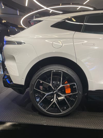 The media file features a close-up, side-view of a white Aston Martin sports car, rotated 90 degrees counter-clockwise. The car's glossy exterior reflects the surrounding lights, which appear to be a modern indoor lighting system with thin, curved white LED strips. The focus is on the rear wheel and fender area.
The wheel is large, black, and multi-spoke with a polished silver rim. Prominently visible are the bright orange brake calipers, which are branded with "ASTON MARTIN" in black lettering. The tire has "PIRELLI" written on its sidewall. The car is parked on a dark, textured floor with a diamond plate pattern, commonly found in showrooms or garages.
To the left of the car, a black metal structure with the curved LED lights is visible, creating an abstract and futuristic ambiance. The lower part of the car shows a sleek black diffuser with hints of blue light emanating from underneath, suggesting internal lighting. The scene is indoors, well-lit, and appears to be a professional automotive showroom or exhibition. There are no people or animals visible. The overall impression is one of luxury, performance, and modern design. The image likely depicts the car displayed in a high-end car dealership or at an automotive event in Doha, Qatar, given the location context.