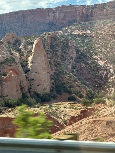 The image shows a mountain landscape. The mountains are tall and rugged, with layers of rock visible. The slopes are covered in sparse vegetation, including trees and bushes. There are some power lines running horizontally across the frame. The foreground is blurry and out of focus, suggesting the photo was taken from a moving vehicle. The sky is partly cloudy. The location is identified as Kayenta, United States of America.