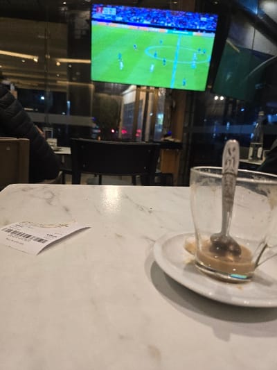 A marble-patterned table with a used cup, saucer, and spoon sits in front of a blurred backdrop of a sports bar showing a soccer game on a large screen. The lighting is dim, suggesting an evening setting. A partially visible check rests on the table's edge. The focus is on the remnants of a drink, with the background offering context.