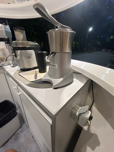 This is a medium shot, vertically oriented image showing a food slicing machine, likely a commercial-grade meat slicer, positioned on a white surface outdoors. The machine is primarily made of brushed stainless steel with black accents. It features a prominent circular blade covered by a safety guard, a handle for moving the food carriage, and a dispensing chute. Text on the machine reads "gmgastro".
In the background, through the opening of what appears to be a food stall or kiosk, a streetlamp illuminates trees and buildings against a dark night sky. The scene is lit by the streetlamp and some interior lighting from the stall, creating shadows and highlighting the reflective surfaces of the machine. To the right of the slicer, a power strip with multiple plugs is visible, indicating the machine is plugged in and ready for use, though no activity is actively occurring. The setting appears to be outdoors at night, possibly in a public area or marketplace in Nova Zagora, Bulgaria, given the location context. The overall mood is neutral, focused on the equipment and the immediate environment.