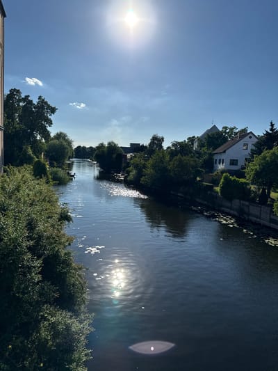 The image depicts a serene canal scene in Rathenow, Germany, bathed in the bright light of a sunny day. The sun is high in the sky, casting a strong glare on the water's surface and creating sparkling reflections. Lush green trees line the banks of the canal, with one side featuring a charming white house nestled amongst the foliage. Lily pads dot the water near the bank, adding to the natural beauty of the setting. In the middle of the canal, a small black boat with a person rowing is visible, adding a touch of human presence to the tranquil environment. The sky is a clear, vibrant blue, with a few scattered white clouds. The overall atmosphere is peaceful and idyllic, suggesting a warm and pleasant day.