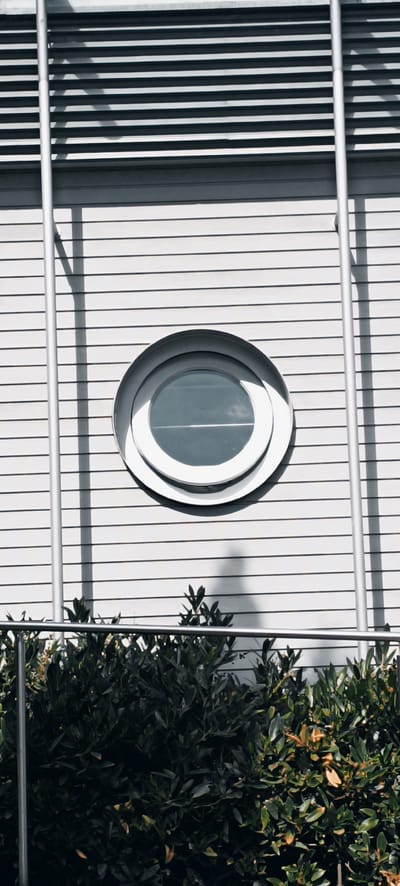 The image shows a close-up of a modern building exterior, likely in the city of Fellbach, Germany, given the context. The facade is composed of horizontal white slats and features a prominent, circular, porthole-style window with a silver frame. To the left of the window, two vertical silver poles run up the building. A shadow of a pole is cast on the wall to the left of the window. In the foreground, a dark green hedge with some brown leaves is visible, partially obscured by a silver railing. The lighting suggests it is daytime, with sunlight creating shadows on the building's surface. No people are visible in the image.