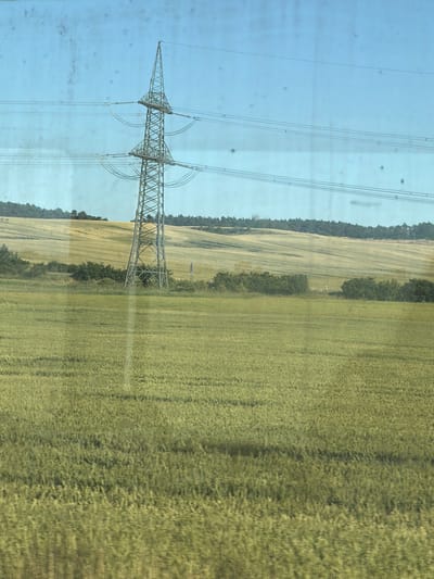 This is a photograph of a power line pylon in a rural landscape. The pylon is made of metal lattice and is supporting multiple electrical wires. It is positioned to the left of the frame, towering over a vast, green field that occupies the majority of the right side of the image. The field appears to be a crop, possibly wheat or barley, with its stalks bending in the breeze.

In the background, a line of trees forms a dark green border along the edge of the field. Beyond the trees, the landscape slopes gently upwards, suggesting rolling hills or a gradual elevation change. The sky is a clear, pale blue, with a few faint, dark specks that could be birds or other small airborne objects.

The image seems to have been taken from a moving vehicle, as suggested by the slight blur and the reflections on what appears to be a window. This motion blur gives the image a dynamic feel, emphasizing the movement of the landscape. The lighting indicates it is daytime, likely late morning or early afternoon, with the sun casting a soft light on the scene.

The overall impression is one of a serene, pastoral setting, highlighting the juxtaposition of industrial infrastructure with the natural environment. The scale of the power line pylon against the expansive field emphasizes its imposing presence in the landscape. There is no visible text in the image.