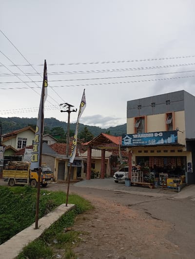 A street scene in Kutawaringin, Indonesia, with a small truck parked on the left side of the road. The truck is loaded with wooden planks. A small shop is visible on the right side of the road with a sign that reads "Warung 8-O" and "Internet - Warung Cafe's LPG". The shop is selling various goods, including bottled drinks. A car is parked in front of the shop, and a few people can be seen inside and outside the shop. The scene is set against a backdrop of lush greenery, and a grey sky. The street is made of gravel and dirt and is lined with foliage on the left. The environment features power lines, and a utility pole on the left. Two banners are visible. The image was captured with a cell phone and does not have a high image resolution.