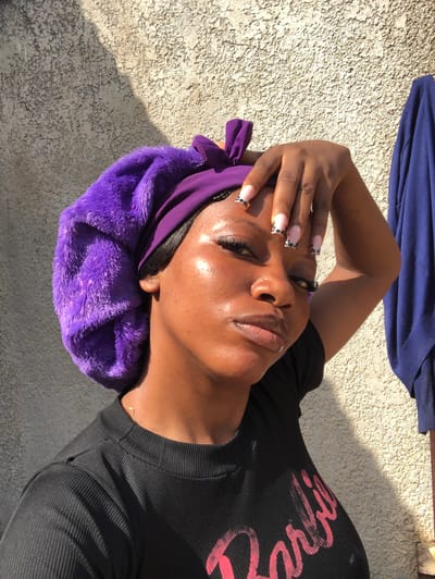 A woman with dark skin is lying down, her head resting on a fluffy purple bonnet. She is wearing a black ribbed t-shirt with pink lettering that reads "Bossy" across the chest. Her eyes are closed, and her hand is resting on her forehead. The lighting suggests it is daytime, possibly with direct sunlight casting shadows on the textured surface she is lying on, which appears to be concrete. Her nails are long and painted with a French manicure featuring black polka dots.