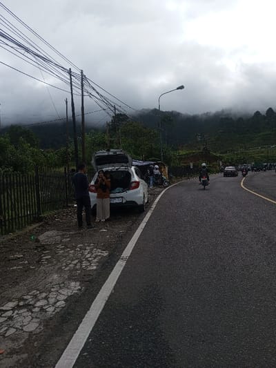 A white car is parked with its trunk open next to a cracked sidewalk and a black metal fence. Two people stand beside the car, while others are gathered nearby. A paved road curves to the right, with a motorcycle and other vehicles driving on it. Overhead, a dense network of power lines stretches across the sky. The background shows a cloudy sky and green hills partially obscured by fog.