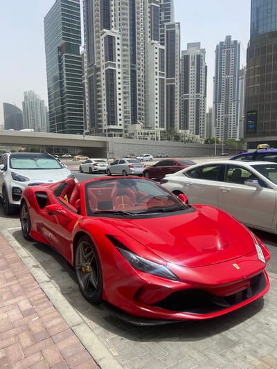 The image shows a street in Business Bay, United Arab Emirates, lined with various parked cars. The foremost car is a red Ferrari convertible. Behind the Ferrari are other cars, including white sedans and other vehicles, parked in a line along the street. In the background, numerous tall buildings and skyscrapers, characteristic of the Business Bay skyline, rise into a hazy sky. The photograph appears to be taken during the day, with sunlight casting shadows on the brick-paved sidewalk where the cars are parked.
