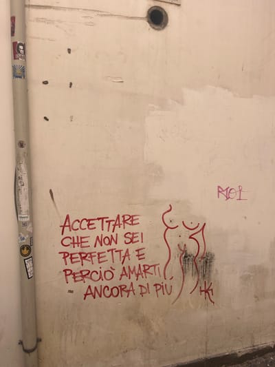 The image shows a section of a wall with graffiti and stickers, located in Salerno, Italy. The wall is a light, weathered color and appears to be outdoors.

On the left side of the image, a vertical pipe is attached to the wall, covered in various stickers. Some of these stickers are faded and difficult to discern, but one appears to be an image of a man with dark hair. Another sticker shows a sunburst design.

The central and right portions of the wall feature red graffiti. The text reads "ACCETTARE CHE NON SEI PERFETTA E PERCIO AMARTI ANCORA DI PIU". This translates from Italian to "ACCEPT THAT YOU ARE NOT PERFECT AND THEREFORE LOVE YOURSELF EVEN MORE." To the right of this text, a stylized outline of a female torso and breasts is drawn in red. Below the torso, a small, abstract symbol or tag is also present in red.

In the upper right quadrant of the image, another graffiti tag in pink reads "RIOI".

The wall shows signs of wear and tear, with some discoloration and small dark marks. The bottom edge of the image reveals a darker, possibly wet or dirty strip along the base of the wall, indicating the ground level. A circular opening, perhaps a pipe outlet or vent, is visible in the upper part of the wall.
