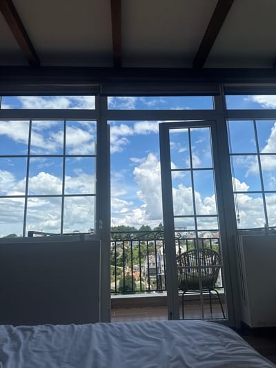 The image depicts a view from indoors, looking out through large, multi-paned windows and an open door onto a balcony and the cityscape of Lam Vien - Da Lat, Vietnam. The interior foreground is dominated by a rumpled white duvet on a bed. The windows themselves are framed with dark material, and the ceiling features exposed wooden beams.
Beyond the glass, a balcony is visible, furnished with a single wicker chair. The balcony railing is made of black wrought iron with a decorative scroll pattern. The exterior view reveals a hillside town with buildings of various sizes and colors, nestled among lush green trees. The sky is a vibrant blue, dotted with numerous fluffy white clouds. The overall impression is one of a bright, possibly sunny day with a pleasant, somewhat humid atmosphere. The scene suggests a domestic setting, possibly a hotel room or a residence, offering a scenic outlook over the city.
