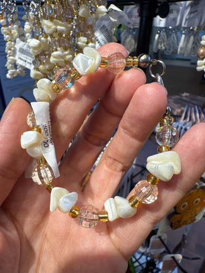 A light-skinned hand is centrally positioned, holding a multi-beaded bracelet horizontally in what appears to be a souvenir shop. The bracelet consists of irregular, polished white shell fragments, clear faceted spherical beads, and small gold-colored spacer beads, connected by a silver-toned metal clasp mechanism. A partially legible white price tag, displaying "200.00" and "Pcs", is attached to the bracelet. The background is out of focus, revealing numerous similar shell and bead jewelry items hanging on various display racks. A black metal display stand and a blurry, light-brown cartoon-like item are also visible in the background. The scene depicts a retail transaction or display of artisanal goods in a commercial establishment in Panglao, Philippines.