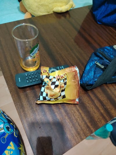 The scene is observed at an unspecified indoor location within the city of Kovachitsa, Bulgaria. A dark brown, wood-grain patterned surface, likely a table or desk, occupies the majority of the frame. On this surface, a clear glass tumbler with a yellow base, bearing a "QUEEN" brand logo with orange and blueberry graphics, stands empty with residual liquid at the bottom. Directly to its right, a black "PRIMA" branded remote control lies horizontally. Partially overlapping the remote and positioned further right is a crumpled yellow, white, and black snack bag, featuring a checkerboard pattern, the text "РУЧКИ" (RUCHKI), and cartoon illustrations resembling chicken drumsticks. To the right of the snack bag, a small, dark blue zippered pouch with a light blue tree-like pattern and a black strap is visible. In the upper left background, a portion of a yellow plush toy rests on a light blue fabric surface. In the lower left, blue fabric with a light blue patterned design is partially visible. In the lower right, the turquoise cap and upper body of a bottle are visible on a lighter surface.
