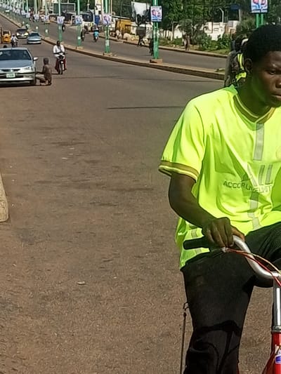 In Garko, Nigeria, on a paved road, a young Black male, wearing a neon yellow t-shirt and black pants, rides a bicycle. He is positioned in the foreground, facing away from the camera, with his hands on the handlebars. Behind him, a silver car is parked on the side of the road. Further in the background, several other vehicles, including motorcycles and cars, are visible on the road, along with pedestrians. The environment appears to be an urban setting with buildings and trees lining the street.