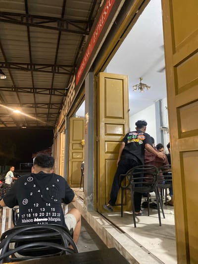 A nighttime exterior shot captures a dimly lit coffee shop in Keudematangpanyang, Indonesia. A man in a black t-shirt with a numerical design sits at a black plastic chair, facing away from the camera. He wears light-colored shorts. To his right, two men are visible through an open doorway. One, wearing a plaid shirt, is being touched on the shoulder by another man in a black t-shirt adorned with colorful graphics. This second man is facing away from the camera, leaning into the doorway. The coffee shop features an outdoor seating area with metal framework and a corrugated metal roof, illuminated by a single hanging lamp. The doorway is flanked by wooden doors, and the interior shows a chandelier and a ceiling fan. The ground is tiled. In the background, a person on a motorcycle is partially visible. The overall impression is of a casual evening gathering at a local establishment.