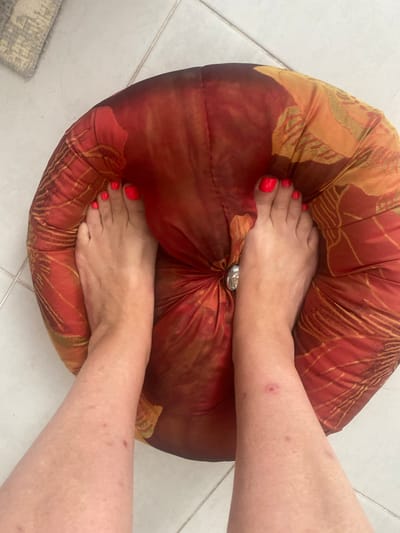 The image shows a top-down view of a person's feet and lower legs resting on a round, red and orange patterned cushion. The toenails are painted a bright neon red. The person's skin appears to have some small red marks or blemishes. The cushion is placed on a tiled floor. A grey and white patterned rug is visible in the upper left corner of the image. The scene is indoors. The location of Novi Petrivtsi, Ukraine, is not directly depicted or implied by the visual elements in the image.