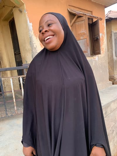 A woman wearing a black hijab smiles broadly. She is standing outdoors in front of a building with a weathered, light brown exterior. There's a window with closed shutters visible above her. The setting appears to be a residential area. A metal fence-like structure is at the bottom left corner. The image is oriented vertically.