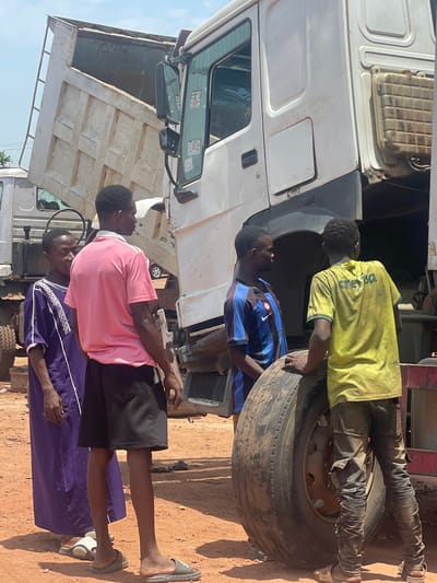 The image depicts a group of four African men working on a large, white truck in Kwali, Nigeria. Two men are focused on the truck's tire, one wearing a yellow shirt with "CERESER" written on the back and the other in a blue and black striped shirt. The other two men are standing in the front of the truck, one in a pink shirt and the other in a purple robe. The environment appears to be an outdoor workspace with a dirt ground.