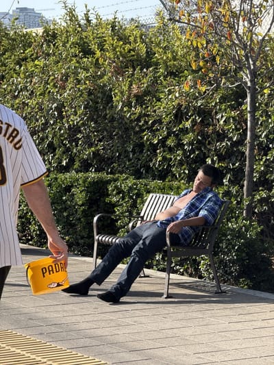 A man is sleeping on a park bench on a paved area near a large hedge and a thin tree. He is wearing black socks and is barefoot, dark denim jeans, and a blue plaid shirt. The shirt is open, and his bare chest is visible. An individual wearing a baseball jersey that is predominantly white with vertical pinstripes and a tan shoulder is holding a yellow bucket with the word "PADRES" in black capital letters. The setting appears to be outdoors in San Diego, United States.