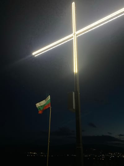 The image shows a night scene in Dolna Grashtitsa, Bulgaria. In the foreground, a brightly lit Christian cross stands atop a post. To the right, a Bulgarian flag is visible, attached to a pole, also illuminated. The background is dark, with some indistinct lights suggesting a town or city.