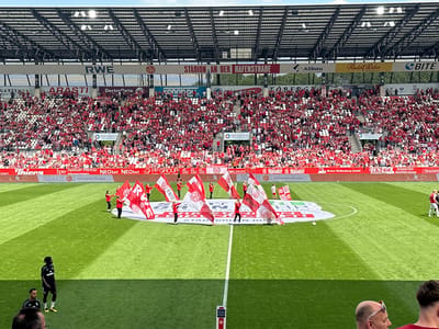 The image depicts a soccer game at Stadion an der Hafenstraße in Essen, Germany. The stadium is filled with spectators dressed predominantly in red. On the field, a group of people are performing, holding large red and white flags with the logo of the team, Rot-Weiss Essen. The stadium has a roof structure with signage for various sponsors, including RWE, Allbau, Rudolf Weber, and Bite. In the foreground, two individuals in black and yellow uniforms are standing on the field. A sign with "MESSE ESSEN" is planted on the field.