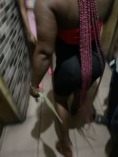 In Alaiyabiagba, Nigeria, a partial interior scene depicts two individuals within what appears to be a tiled corridor or room. The primary subject, seen from behind, is a person with dark skin, wearing a red strapless top, black form-fitting shorts, and long, burgundy box braids extending down their back. A gold-colored bracelet is visible on their right wrist. This individual holds a single, elongated light green leaf in their right hand and wears brown low-heeled footwear on their right foot, appearing to be in motion. To the immediate left of this subject, a wall is tiled with light-colored, rectangular tiles arranged in a diagonal pattern. Further in the background, a second, highly blurred individual is partially visible, wearing a light pink garment and a multi-colored (rainbow-striped) top. The floor is light-colored and tiled. The scene is dimly lit, casting shadows.