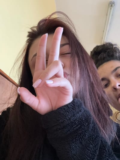 The image shows two young females captured indoors within the city of Cherven Bryag, Bulgaria. The foreground subject, positioned left, has long, dark reddish-brown hair and wears a black, textured fleece garment. Her left hand is raised, partially obscuring her face, with the index, middle, and ring fingers extended vertically. Her fingernails exhibit a light pinkish-white manicure. Her visible right eye is narrowed, and her mouth is puckered. Behind her, in the upper right, a second female subject is partially visible, featuring dark hair styled in an updo. She is seen extending her tongue. Her visible left eye is directed towards the camera. The immediate background features a plain light-colored wall on the left, and a white ceiling with a single fluorescent light fixture in the upper right. A dark wooden element is visible on the far-left edge of the frame.