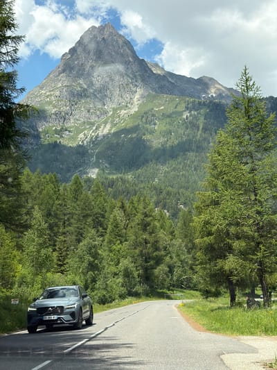 The image depicts a mountainous landscape with a winding road in the foreground. A grey Volvo XC60 SUV is driving on the road, with its front facing the camera. The car is positioned on the left side of the road as it curves to the right. The road is asphalt and bordered by green grass and trees.

Behind the road, a dense forest of pine trees covers the lower slopes of the mountains. The upper part of the mountains is rocky and barren, with patches of green vegetation and snow visible. The sky is partly cloudy with a mix of blue sky and white clouds. The scene appears to be captured in a natural, outdoor setting, likely a scenic route in a mountainous region.

The location is identified as Vallorcine, France, a commune in the Haute-Savoie department in the Auvergne-Rhône-Alpes region of southeastern France. The presence of the Volvo XC60, a modern SUV, suggests a contemporary setting. The overall impression is one of a serene and picturesque drive through the French Alps.