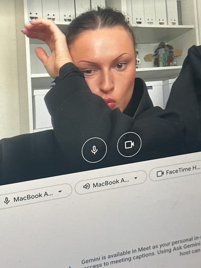 The image depicts a person with dark hair and a black hooded sweatshirt resting their head on their hand. In the background, there is a white shelf with several white drawers or boxes. On the right side of the image, there is a laptop screen displaying FaceTime and Meet options. Text on the screen mentions "MacBook A..." and "Gemini is available in Meet as your personal in-." Above the person's head and to the left is a Star Wars toy. There is no indication of Wealden, United Kingdom, in the image.