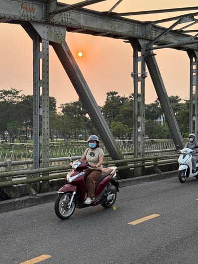 The image captures a scene on a bridge in Phú Xuân Ward, Vietnam, during sunset. An orange sun shines through the bridge’s steel truss structure. Two people are riding motorbikes. One person, wearing a helmet and a surgical mask, is riding a red scooter. Further ahead is another scooter, with two riders, partially visible. Yellow lines mark the road on the bridge. Trees and buildings are visible in the background.
