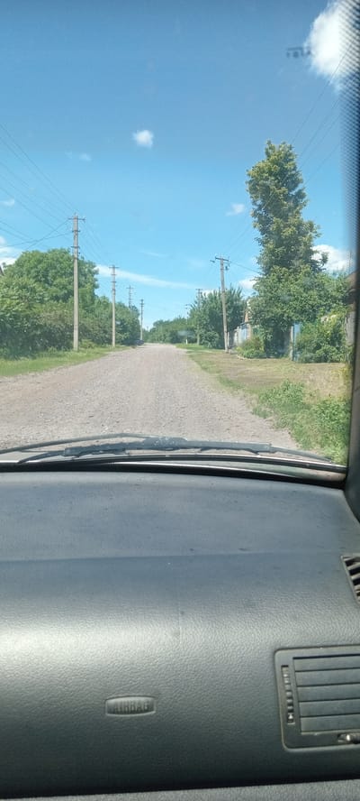 The image shows a view from inside a vehicle, looking out through the windshield at a dirt road in a rural area. The road is flanked by trees and power lines. The sky is blue with a few scattered clouds. The dashboard of the car is visible in the foreground, with the word "AIRBAG" embossed on it. The location is described as Koviahy, Ukraine, suggesting a context of everyday life in a Ukrainian village or town. The scene is peaceful and ordinary, depicting a common sight of travel through a rural landscape.