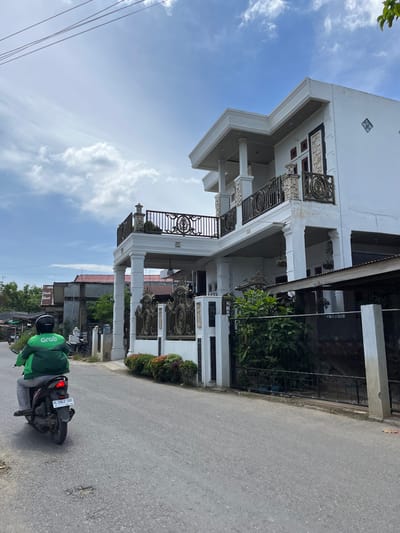 This is a medium shot, eye-level photograph taken from a street in Langsa, Indonesia. The scene depicts a white, two-story house with decorative railings on its balconies and a tall, ornate gate. In the foreground, a person wearing a green Grab jacket and helmet rides a black motorcycle away from the camera. The motorcycle has a license plate visible, reading "BL 5942 DBE". The street appears to be paved with asphalt and shows some signs of wear, including potholes. The sky is blue with scattered clouds. The surrounding environment includes other houses and vegetation.