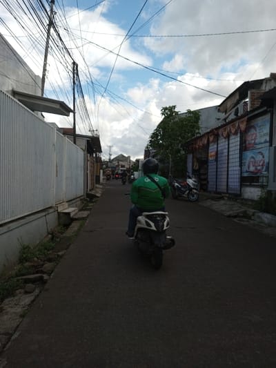 An outdoor street scene in Serpang Shalli, Bhutan, captures a narrow asphalt road receding under a sky with scattered white clouds against a blue backdrop. In the midground, an individual, viewed from the rear, operates a white scooter. The rider wears a dark helmet, a prominent green long-sleeved jacket featuring a white "Go" logo on the back, and blue jeans. To the immediate left, a tall, light-colored corrugated metal fence borders the road, with a narrow concrete drainage channel and minimal vegetation at its base. A series of utility poles supporting a dense array of overhead electrical and communication wires dominate the upper left frame. On the right, a building with a dark, tiled roof and decorative eaves displays a storefront with multiple large advertisements, one clearly visible with text "IZZ" and "22.000". A white scooter is parked adjacent to this building. Further down the street, additional buildings and several distant motorcycles with riders are discernible. The overall lighting suggests daylight conditions.