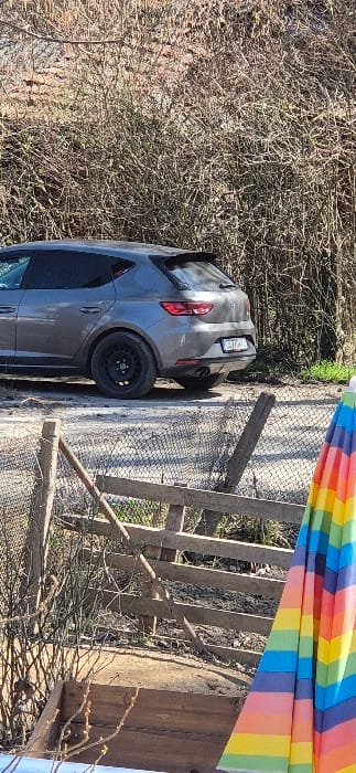 The scene is located in Tompsan, Bulgaria.

A grey, five-door SEAT Leon hatchback is parked on a gravel shoulder to the left of a sparse, deciduous brush line. The vehicle's rear is visible, featuring a single exhaust pipe on the left, dark-tinted rear windows, and black steel wheels. The Bulgarian license plate "CB T104KL" is affixed to the rear. The background consists of dense, bare deciduous vegetation under bright sunlight. In the foreground, a rudimentary wooden fence with wire mesh runs diagonally across the lower half of the frame. To the immediate right, a partially folded, multi-colored (rainbow-striped) beach umbrella stands vertically. Below the fence, various wooden planks and ground debris are visible. No individuals are present in the image.