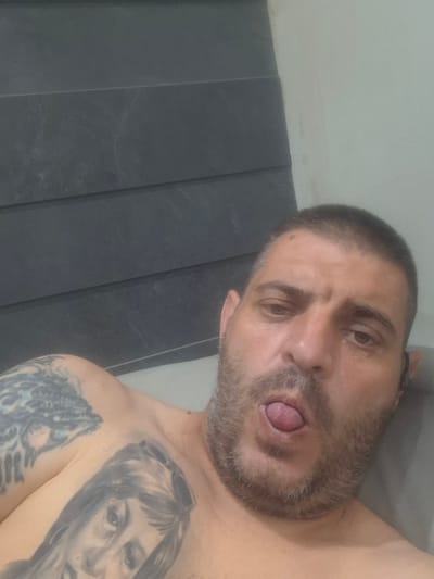 An adult male is depicted in a close-up, slightly upward-angled shot, shirtless from the chest up. He has short, dark hair, a full beard mixed with grey, and appears to be in his 30s or 40s. His eyes are partially closed or squinted, and his mouth is open with his tongue extended. A dark earbud is visible in his right ear. He displays multiple tattoos: a large, dark, portrait-style tattoo of a woman's face is visible on his left chest, and an intricate, dark, abstract or organic design covers his left upper arm and shoulder. The background features a dark grey wall with horizontal, subtly textured panels to the left, and a plain light grey wall to the right. The scene is located in the city of Sopot, Bulgaria.