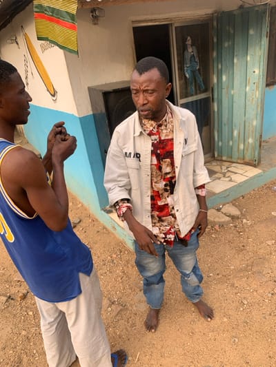 This image depicts two adult males interacting outdoors in Okene, Nigeria, against the backdrop of a building.
The man on the left, facing right, wears a blue basketball jersey with yellow accents and light-colored pants, completed by blue flip-flops. His hands are clasped at chest height, and his mouth is slightly open as if speaking or mid-sentence.
The man on the right, facing left, has a distinct V-shaped hairline. He wears an unbuttoned light beige over-shirt with "MIR" visible on the left chest, layered over a multi-colored, long-sleeved collared shirt with red, white, brown, and black patterns. He wears distressed blue jeans and is barefoot. A light-colored bracelet adorns his right wrist, and a small, dark object (possibly a phone) is held in his left hand. His facial expression is somewhat pensive, with furrowed brows, and he appears to be looking downwards and to his left, seemingly listening or reflecting.
The men stand on a dirt and gravel surface. Behind them, a building wall features a light-colored base with a horizontal blue stripe. A mural on the wall depicts a long, yellow, club-like object with black outlines, accompanied by partial text "COC..." and numbers "080808..." below it. Above the mural, a flag with horizontal red, yellow, and green stripes is visible. To the right of the mural, a dark rectangular speaker or utility box is situated next to a doorway. This doorway is fitted with a screen door, behind which a cutout or poster of a person in casual attire is visible. Further right, a light blue rolling metal door or shutter is partially open. The scene is illuminated by natural daylight.