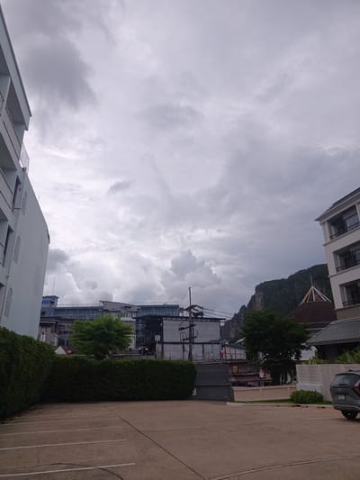 The image shows a parking lot in Ban Ao Nang, Thailand on a cloudy day. The lot is mostly empty, marked with white lines, and appears to be made of concrete. It is bordered by green hedges and buildings of varying heights. On the left is a white building with balconies. In the background, several other buildings can be seen. To the right, a building with a pointed roof, similar to a temple, is visible behind a modern, white-walled structure. A dark gray SUV is parked on the far right. Above the buildings, a rock mountain formation can be seen on the right, with dense clouds covering the sky. Utility poles with wires run across the middle ground. No people are visible in the image.