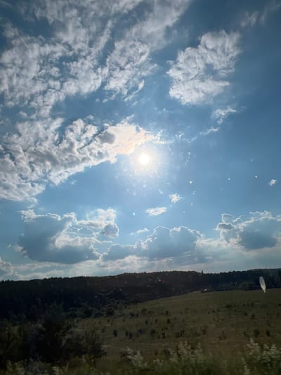 A vast, partly cloudy sky dominates the upper portion of the image. The sun, a bright orb, shines through a gap in the clouds, casting rays and creating lens flare effects. Cumulus clouds, both white and gray, are scattered across the blue expanse. Below the sky, a dark, tree-covered hillside slopes down to a wide, green field dotted with sparse vegetation. The field is illuminated by the sun, with shadows cast by the clouds. The foreground features blurry green foliage, suggesting the photo was taken from a moving vehicle. The scene is a natural landscape, likely in a rural or semi-rural area, possibly near Hrabovets, Ukraine. There are no people or specific man-made structures clearly visible.